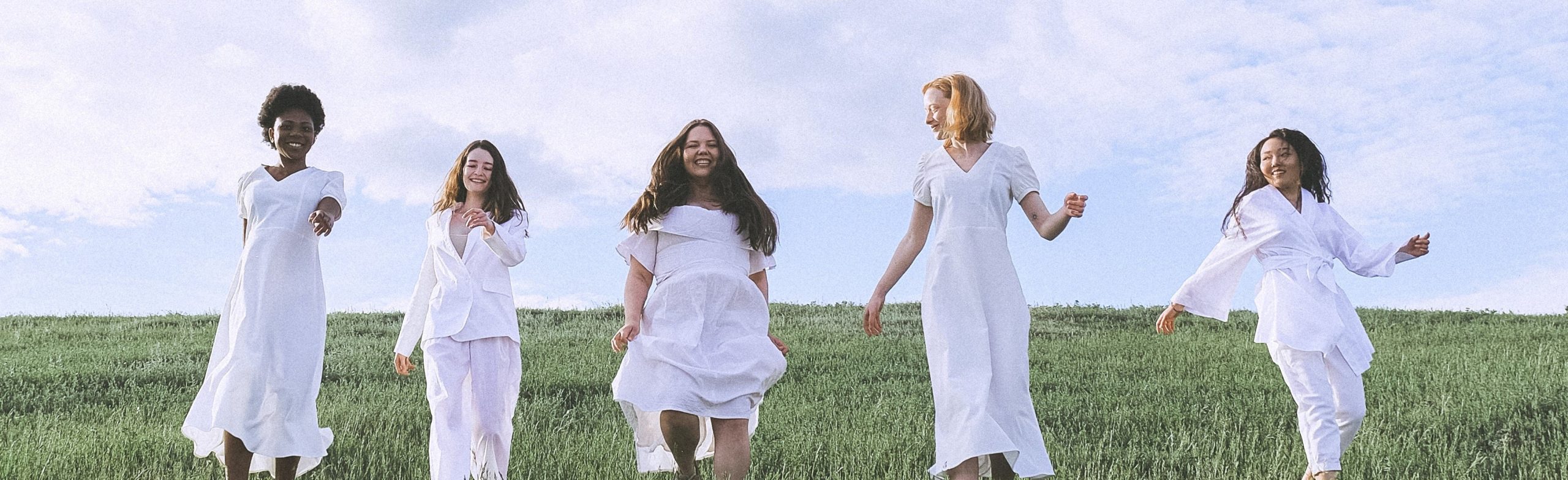 five women of different skin colour and cultural background, all wearing white clothes, walking together across a field of green grass, smiling